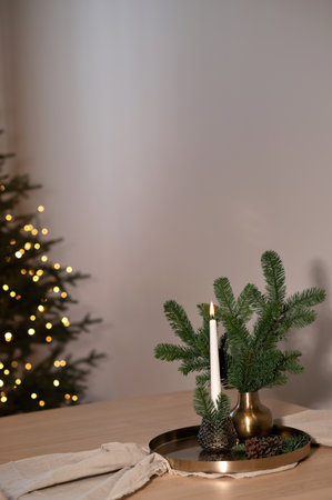 Aesthetic Christmas holiday home interior decoration, candle, vase with tree branches on golden tray on beige wooden table, blurred Christmas tree on background, empty neutral wall copy spaceの写真素材