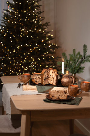 Festive Christmas holiday table setting, wooden table with food, panettone, tea cups, linen napkins, pine branch and candlelight, blurred Christmas tree lights on background, lifestyle home interiorの写真素材