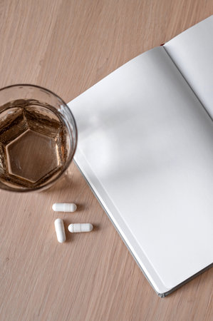 White pills and water glass, open notebook with empty sheet mockup copy space on neutral beige wooden table background, healthy lifestyle habits and wellness concept.の写真素材