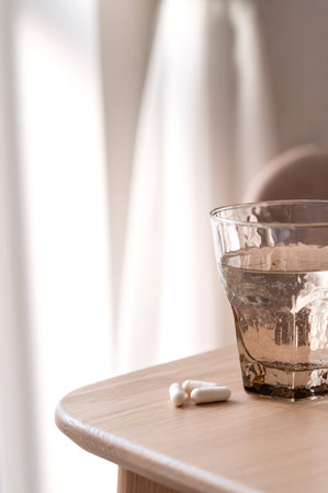 Water glass and white supplement capsules on neutral beige wooden table with soft natural sunlight, healthy morning routine, self care and wellness concept.の写真素材