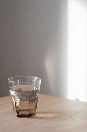 Glass with pure water on neutral oak beige wooden tabletop, soft natural sunlight on empty white...