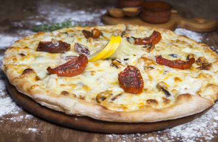 pizza with cheese sun-dried tomatoes seafood and lemon on a brown wooden table with scattered flourの写真素材