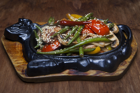 fried pork with grilled vegetables and sesame seeds in an iron plate on a brown tableの写真素材