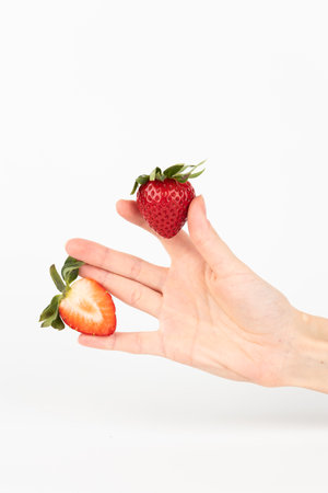 on a white photo background, thin female hands hold a red ripe strawberry with a green tail between the fingers, a cut half of a strawberry, healthy vitamins, seasonal summer fruit, vitamin Cの写真素材