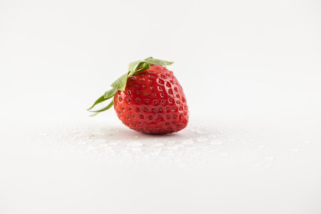 on a white photo background lies one ripe strawberry in the center of the background drops of water healthy vitamins seasonal summer fruit vitamin C green strawberry tail like a crownの写真素材
