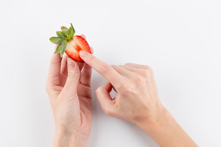 on a white photo background, thin female hands hold a cut strawberry with a finger poking into the pulp of the berry, healthy vitamins, seasonal summer fruit, vitamin Cの写真素材