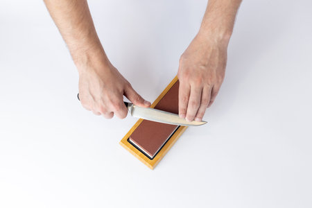 top shot on a white background on a table, man hands sharpen a steel knife protection against cuts, a red medium-hard stone for sharpening knivesの写真素材