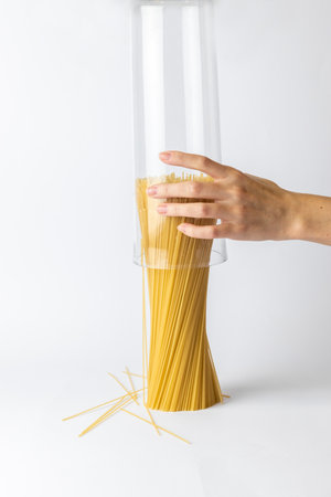 close-up on a white background from the side a woman's hand above the table holds spaghetti with the bottom to the top spaghetti outside the container stands on the table a cylinder of spaghettiの写真素材