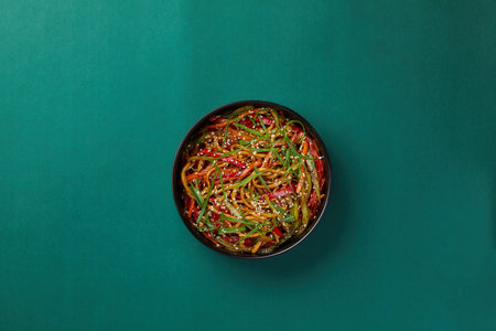 on green background, a black round bowl of rice noodles with striped vegetables sprinkled with sesame seedsの写真素材