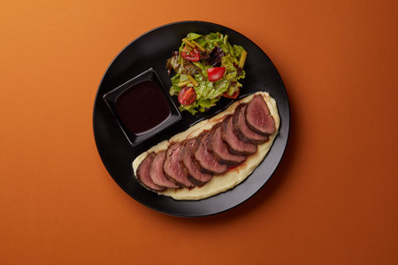 on orange background on a rectangular black plate lies mashed potatoes on which pieces of filet mignon are laid out, served with mushrooms and tomatoの写真素材