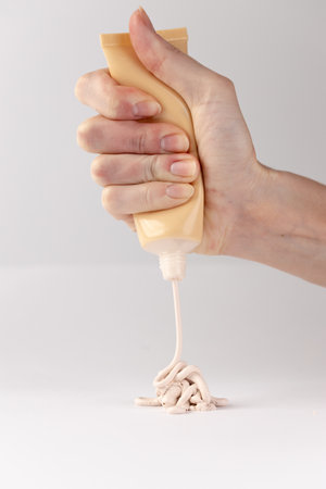 A Close-Up View of a Hand Squeezing a Tube, Dispensing Beige Cream or Paste onto a Clean White Surface for Art, Crafting, or Cosmetic Applicationsの写真素材