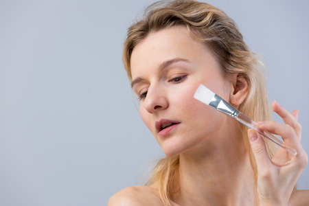Woman with blonde hair applying skincare products using a brush, showcasing a beauty routine focused on facial care and enhancing natural features in a soft lightの写真素材