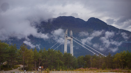 A Majestic Bridge in the Heart of Dramatic Landscape Surrounded by Mountains and Cloudy Skies Reflecting Natures Splendor and Architectural Ingenuityの写真素材