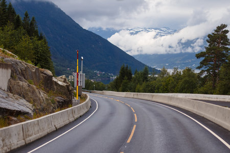 Scenic Road Curving through Mountains with Lush Greenery and Cloudy Sky in Norways Rugged Landscape Captured in a Serene Moment of Natureの写真素材
