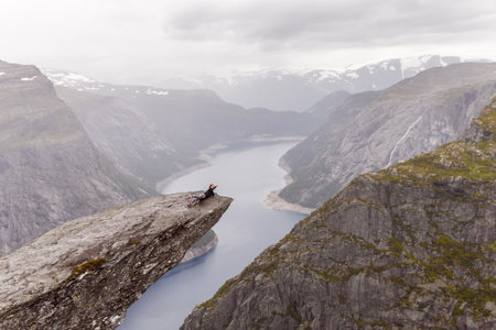 Breathtaking View from a Dramatic Cliff Edge with Person Relaxing in a Stunning Norwegian Landscape Overlooking a Majestic Fjord and Mountains Under a Cloudy Skyの写真素材