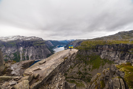 An Adventurous Hiker Waves While Standing on the Edge of a Stunning Cliff with Breathtaking Views of Fjords and Snow-Capped Mountains in Norwayの写真素材
