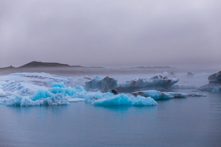 A Tranquil Glacial Landscape Under Cloudy Skies: The Beauty of Icebergs Reflected in Calm Waters with Mysterious Fog Enveloping the Wilderness Beyondの写真素材