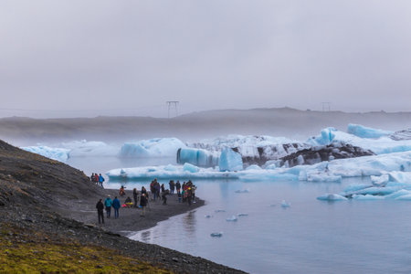 Spectacular Glacier Lagoon with Tourists Amidst Floating Icebergs and Misty Landscape, Offering a...