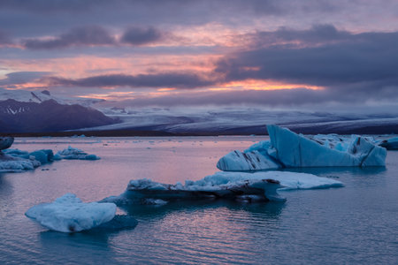 A Stunning View of Icebergs in a Serene Glacier Lagoon Under a Colorful Sunset Sky with Majestic Mountains in the Background During Early Eveningの写真素材