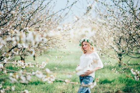 Beautiful young cheerful woman in wreath of flowers on head touching belly while walking in spring tree garden. Beauty People Lifestyle concepts. Happy mom in waiting for baby!の写真素材