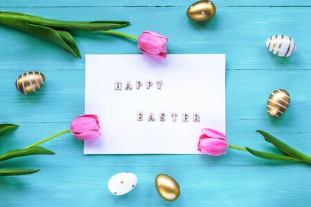 Festive Easter background. White sheet with words Happy Easter on turquoise backdrop with flowers and painted eggs. Top view flat lay.の写真素材