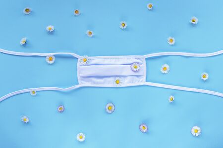 White protective medical mask on blue background with daisy flowers. Covid-19 concept. Top view, flat layの写真素材