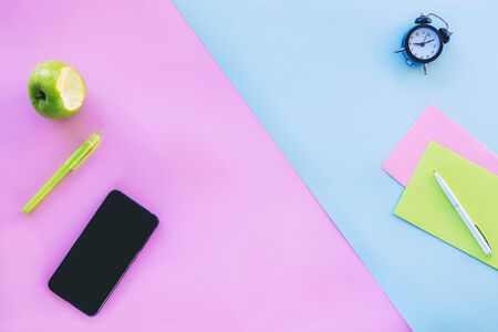 Mobile phone, pen, green apple and office accessories on pink and blue background. Work from home...