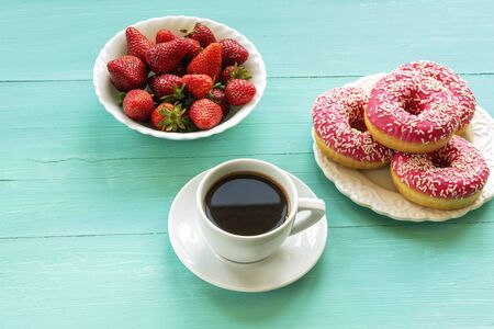 Cup of coffee, bowl with fresh strawberries and white plate with pink baked donuts on turquoise painted wooden table, top view.の写真素材