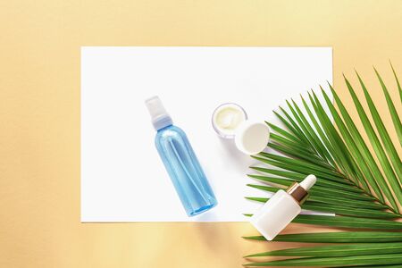 Facial and body care cosmetics bottles on yellow background. Serum, moisturizer, cream jar unbranded blank packages on white paper with palm leaf. Top view, flat lay, copy spaceの写真素材