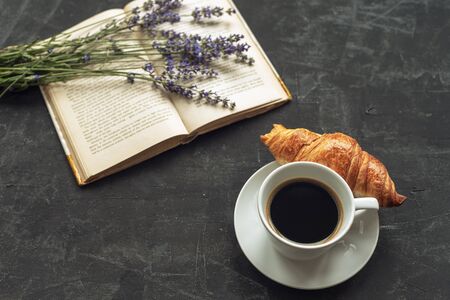 Cup of coffee with croissant, book and lavender bouquet on black table.の写真素材