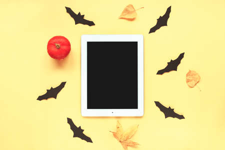 Tablet with blank screen and pumpkin, autumn leaves, handmade black bats on yellow background. Halloween concept. Top view, flat lay.の写真素材