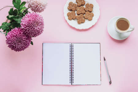 Cozy woman workplace with chrysanthemum flowers, blank notebook, cup of coffee and cookies on pink background. Top view, flat lay, copy space.の写真素材