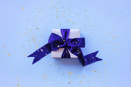 A gift in a box on blue festive background with golden confetti. Top view, flat lay.