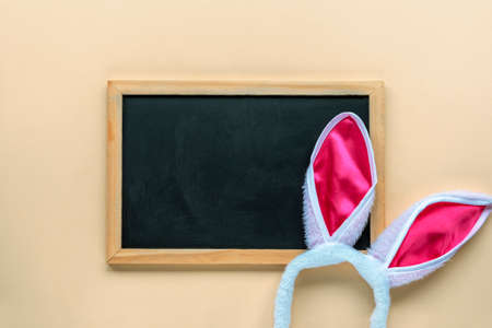 Blank blackboard and bunny ears costume headband on beige background. Easter concept. Top view, flat lay, mock up.の写真素材