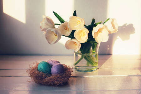 A bouquet of white tulips and a nest with colored eggs on a white wooden table. Easter decorations,...