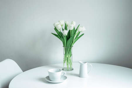 Bouquet of white tulips in a glass vase, a cup and a milk jug on a white table. Morning, breakfast, spring concept.の写真素材