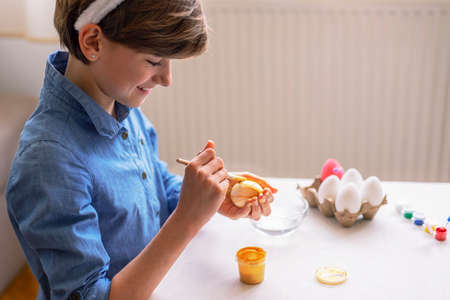 Beautiful nine-year-old girl in blue shirt paints eggs at a white table. Easter concept. Close up.