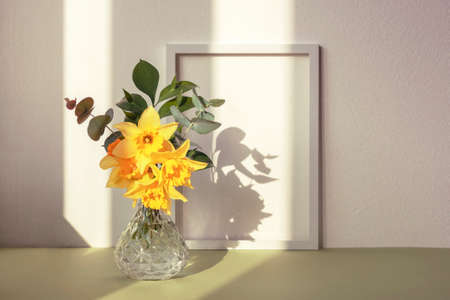 Daffodil bouquet in glass vase and picture frame against white wall in sunlight. Spring concept.の写真素材