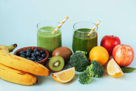 Two different green smoothies in glasses with straws, banana, apples, broccoli, kiwi, lemon and blueberries in a bowl. Healthy ingredients, white backgroundの写真素材