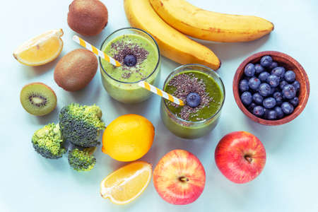 Two different green smoothies in glasses with straws, banana, apples, broccoli, kiwi, lemon and blueberries in a bowl. Healthy ingredients, bright blue backgroundの写真素材