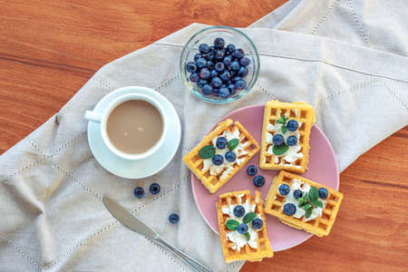 Waffles with cream and blueberries, a cup of coffee on the table on a cloth napkin. Breakfast concept. Top view, flat lay.の写真素材