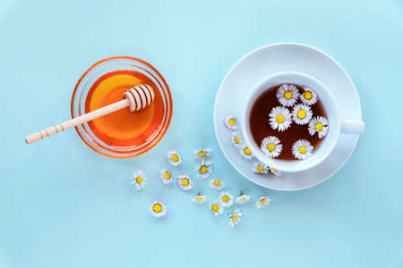 A cup of tea with chamomile and honey in glass bowl with wooden dipper on blue background