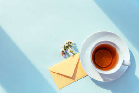 A cup of tea, an envelope and a bouquet of daisies in sunlight on blue background. Top view, flat layの写真素材