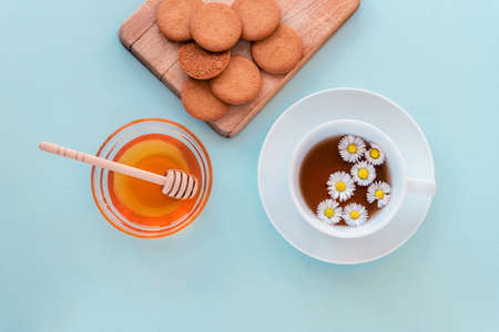 A cup of tea and honey in glass bowl with wooden dipper and cookies on cutting board on blue background. Healthy breakfast. Top view, flat lay.の写真素材
