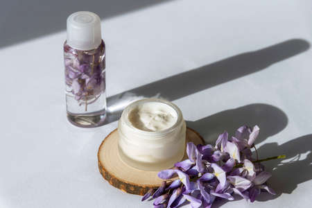 Cream jar and purple wisteria flowers on white table in sunlight. Natural organic cosmetics concept. Long harsh shadows, top view.の写真素材