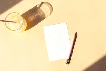Blank sheet on the table, pen and a glass of lemonade with a straw in sunlight. Summer concept. Top view, flat lay, mock upの写真素材