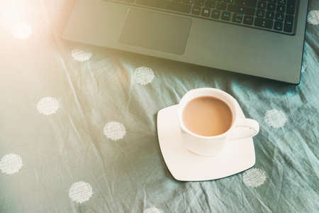 Workspace in bed with laptop and coffee in sunlight. Top view.