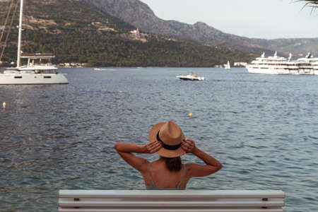 Girl in a straw hat sits on a bench overlooking the sea and the island. Summer vacation concept. Adriatic sea, Croatiaの写真素材