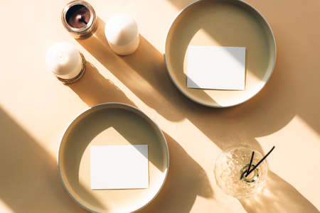 Blank paper sheet cards mockup on beige background in sunlight. Ceramic plates and candles, aroma sticks in glass vase with long shadows. Top view, flat lay.の写真素材