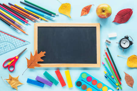 Blackboard and school supplies stationery, colour pencils, paints, scissors, ruler, alarm clock, apple, autumn leaves on blue background. Back to school concept. Top view, flat lay. Mock up.の写真素材